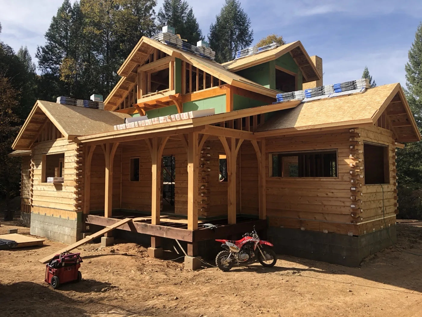 Log home framing with covered porch posts and a dirt bike parked in the forested yard