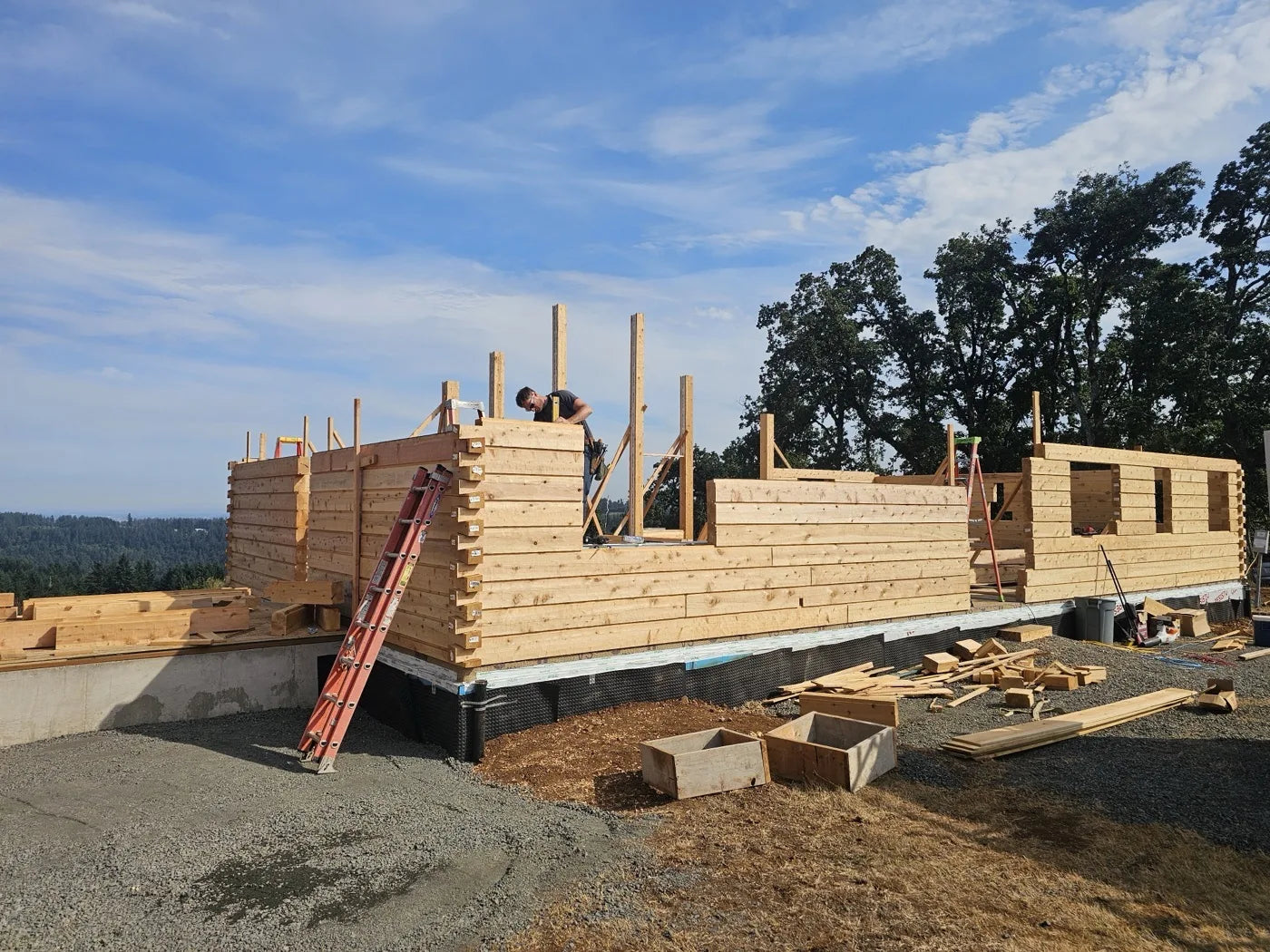 Stacked log walls under construction on a clear day with oak trees and rolling hills