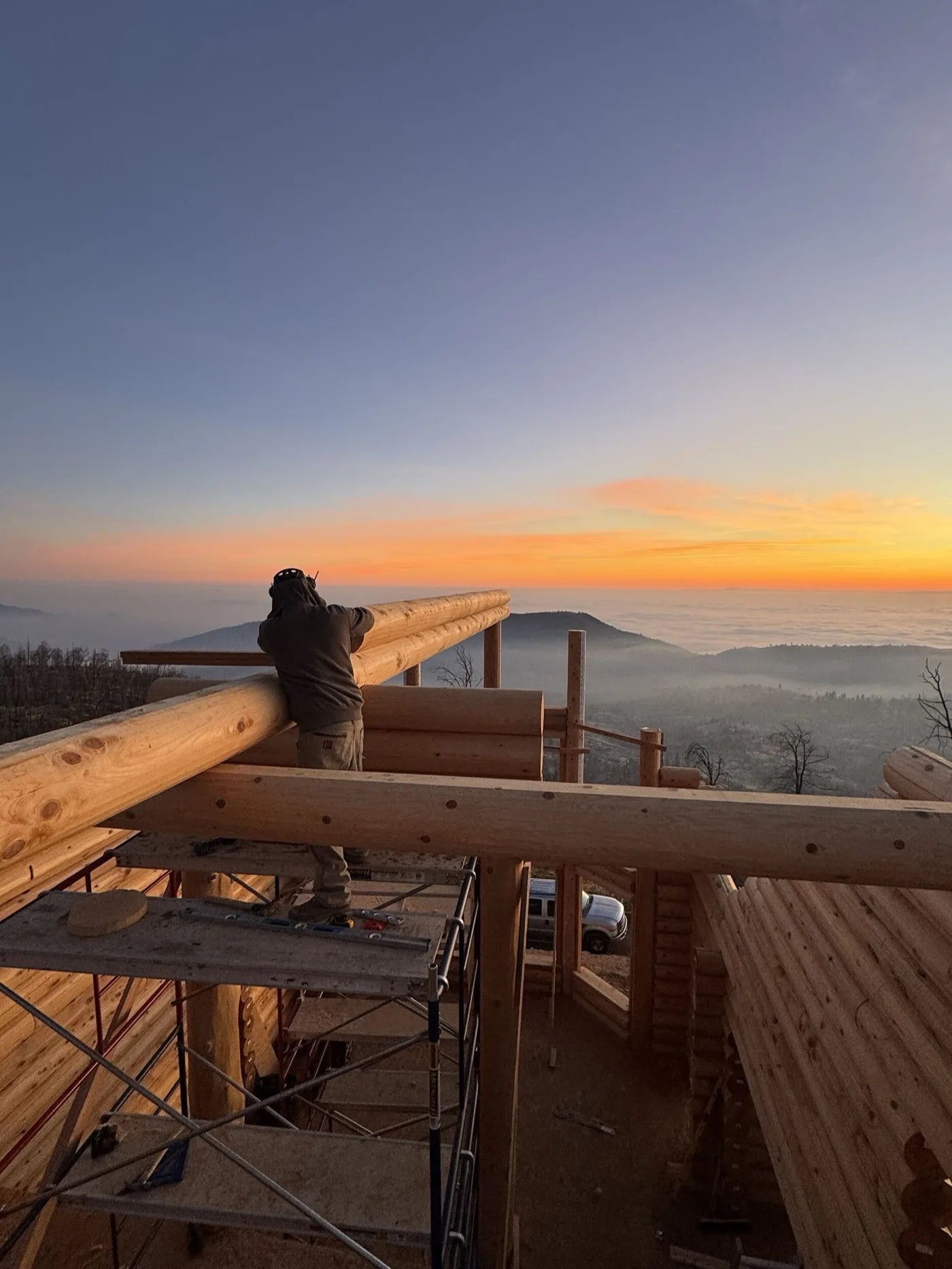 Worker lifting a roof log beam at sunrise above a fog-covered valley