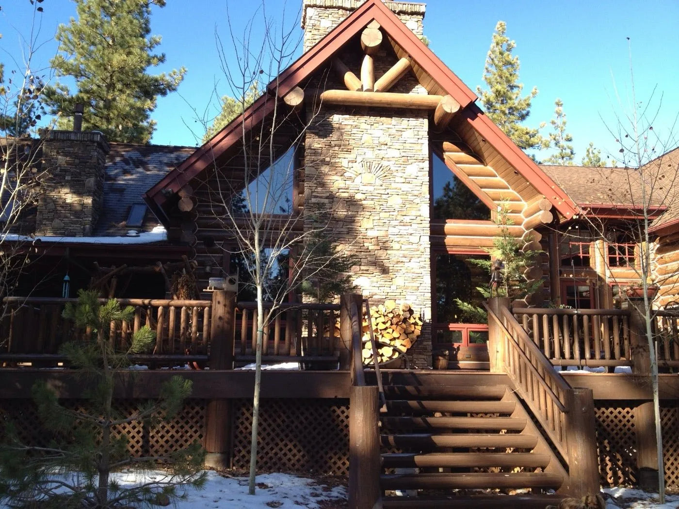 Log home exterior with a red gable roof, stone chimney, and stacked firewood in winter